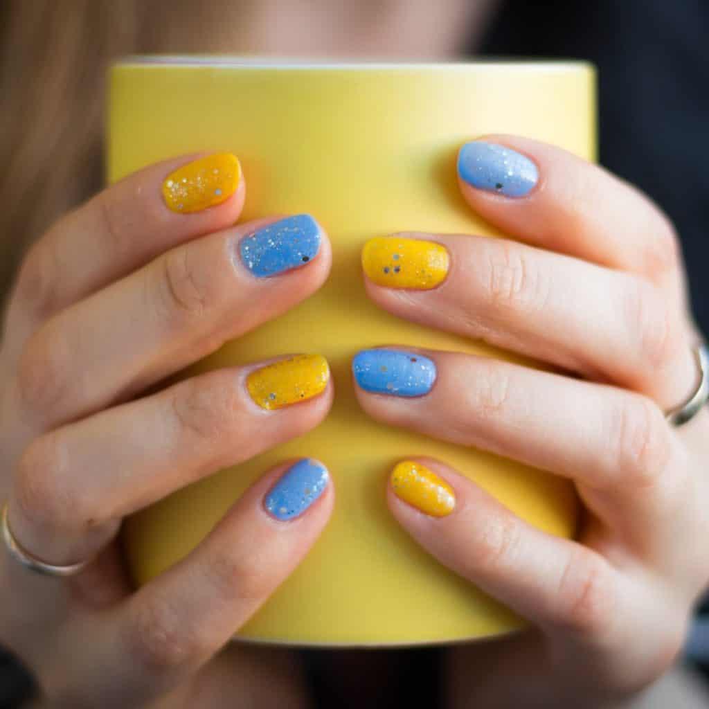 a woman holding a yellow cup flaunting her cute round yellow and blue manicure with glitters