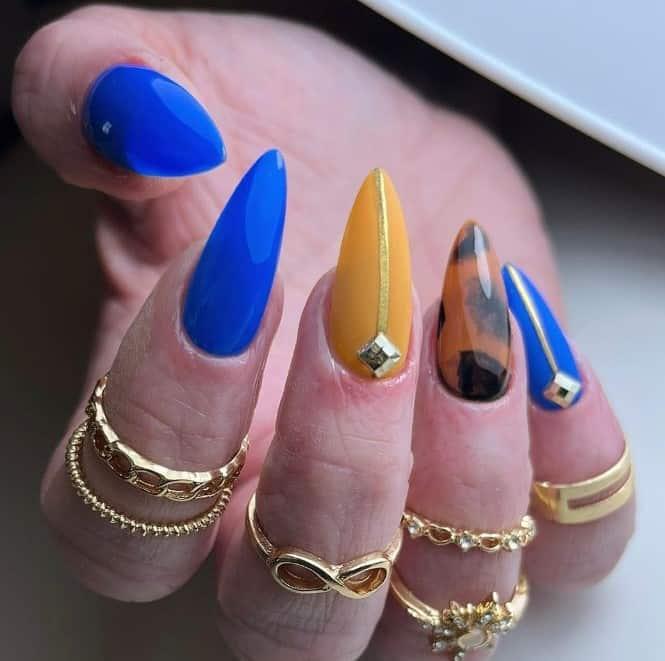 a woman stiletto nailswith glossy bold blue and yellow polish, gold embellishments, and a stunning tortoiseshell accent nail