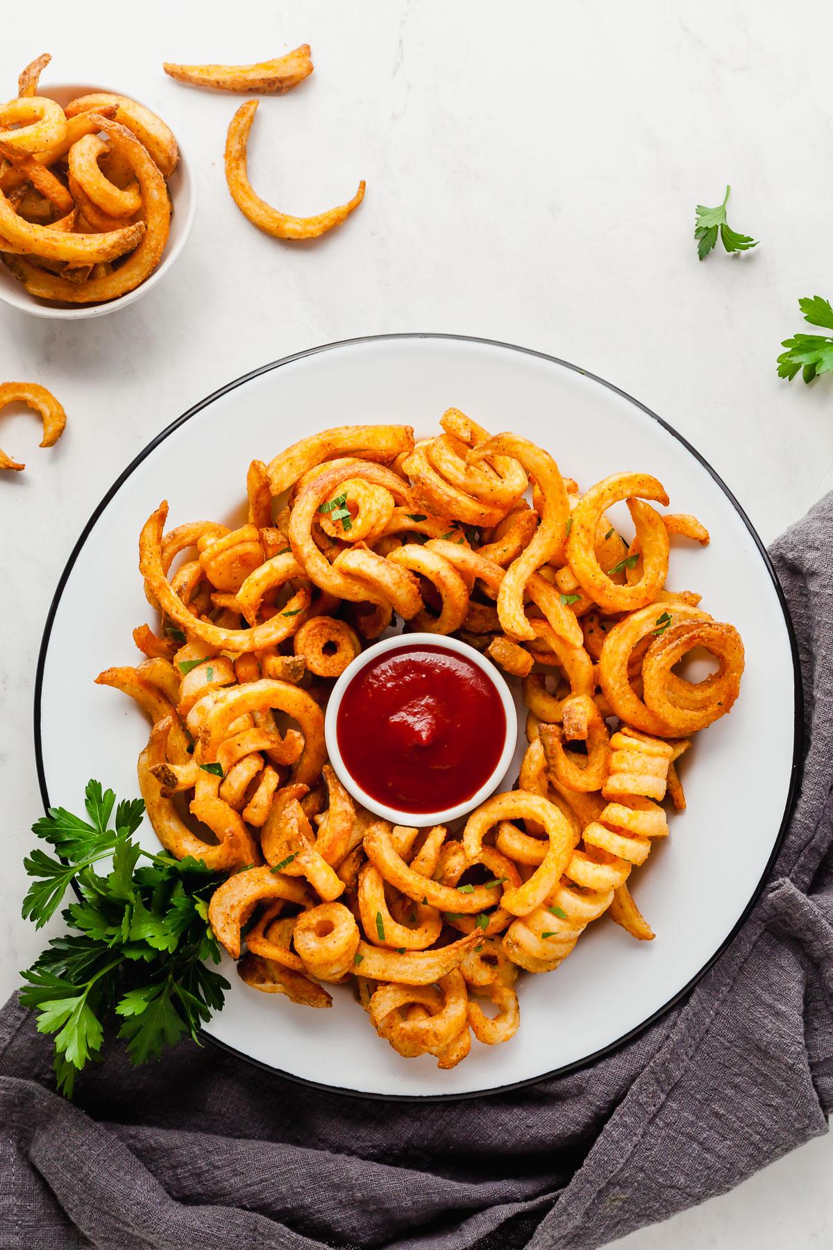 frozen curly fries made in the air fryer on a plate surrounding a bowl of ketchup