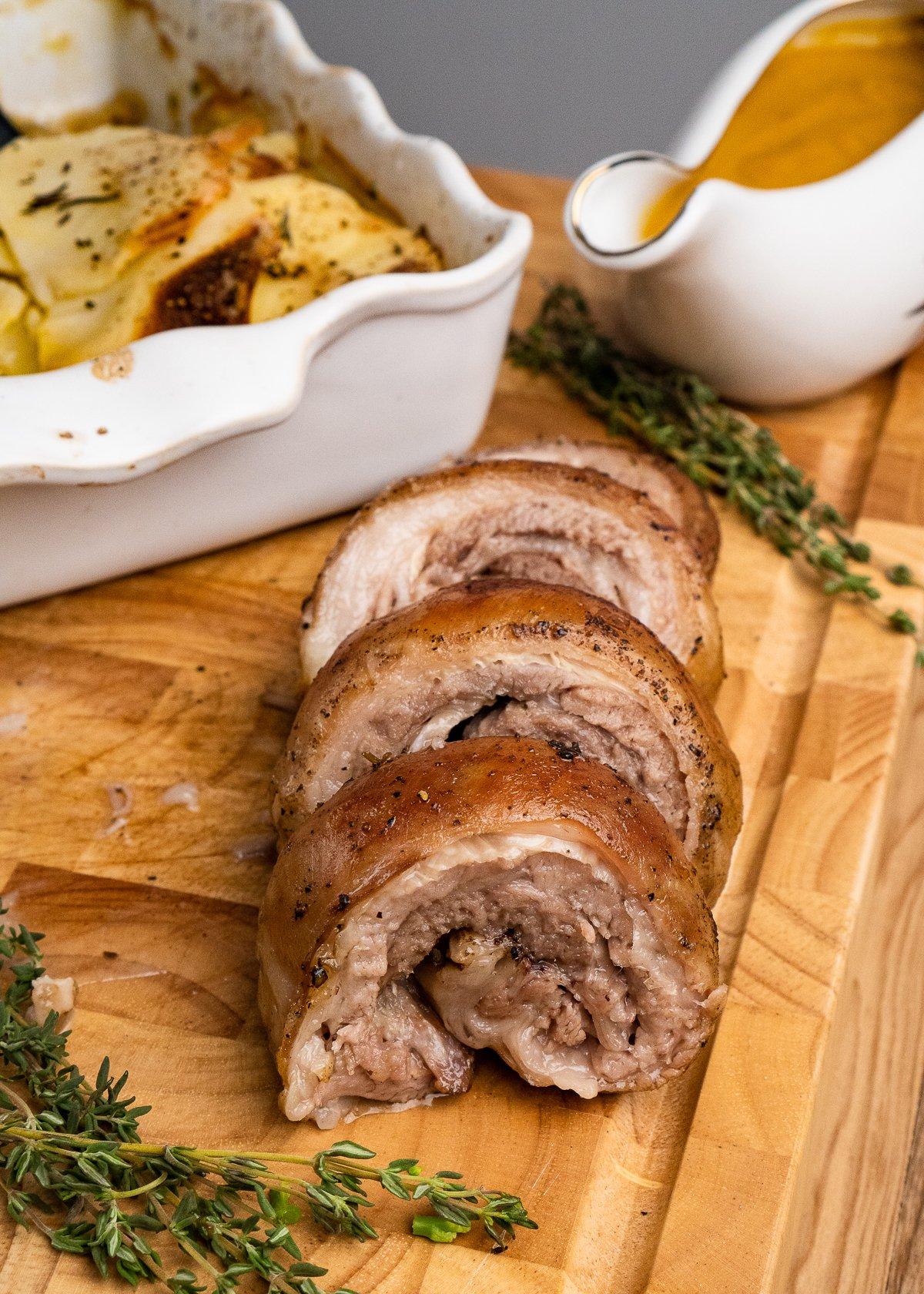 Slices of Lamb Breast with boulangerie potatoes, sprigs of rosemary and a gravy boat filled with lamb sauce in the background