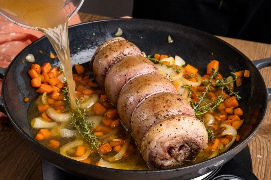Seared lamb breast in a cast iron pan with chopped onion, garlic, and carrot with sprigs of rosemary and pour in lamb stock around the sides