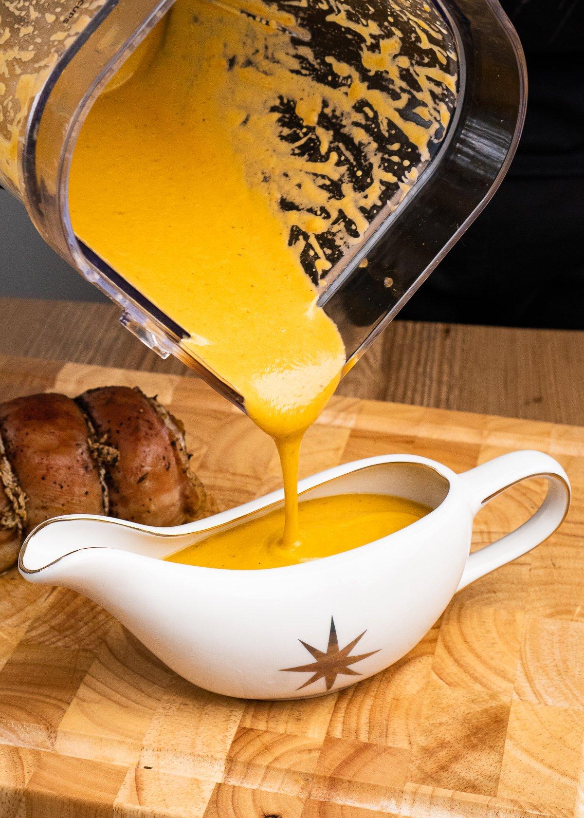 Lamb sauce being poured into a gravy boat from a blender with a lamb breast joint on a wooden chopping board