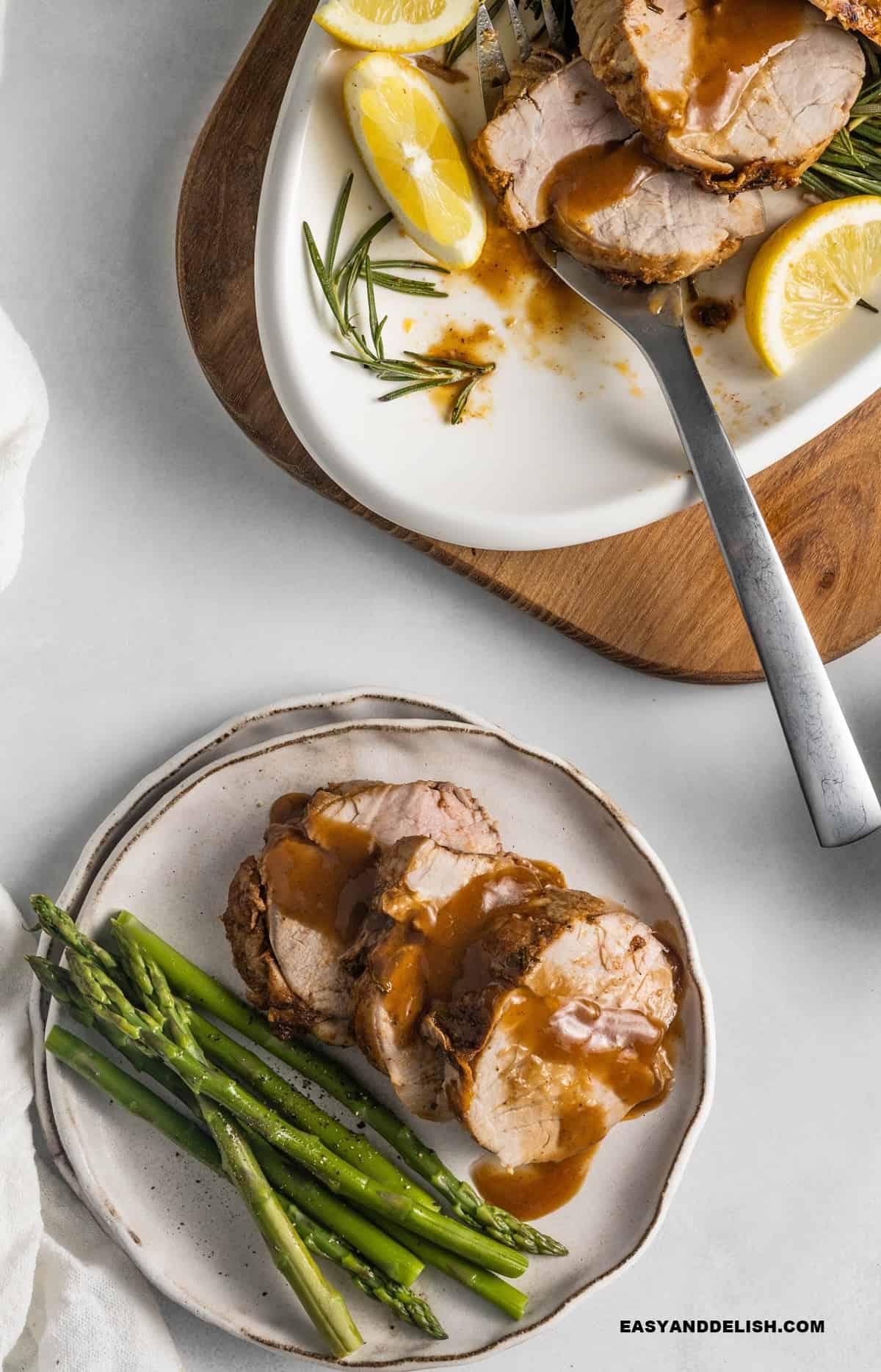 Sliced pork tenderloin with wine sauce on a plate served with asparagus and a platter in the background.