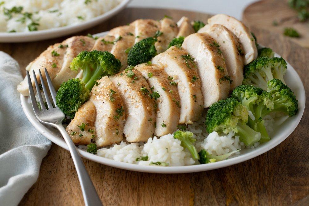 White plate with rice, broccoli and sliced toaster oven chicken breast. Fork and gray napkin on left.