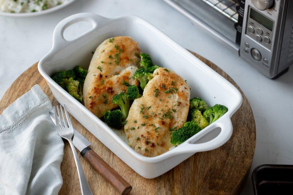 Overhead view of baked chicken breast and broccoli in white baking dish. Round wood board underneath. Gray napkin, knife and fork on left and toaster oven in upper right.