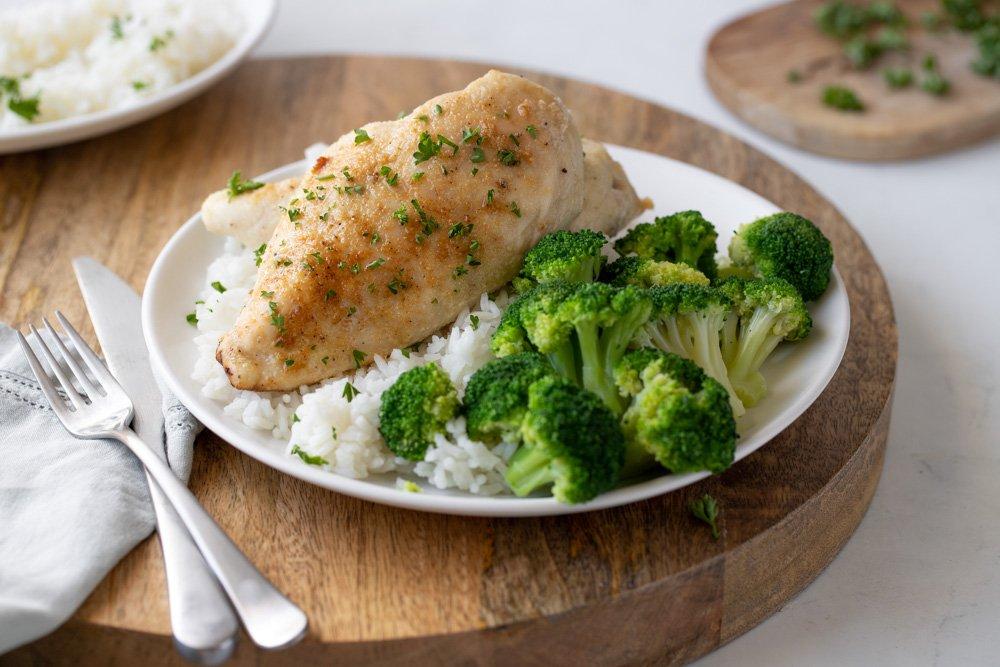 White plate with rice, broccoli and toaster oven chicken breasts. Fork and gray napkin on left. Cutting board with parsley in back right and plate with rice in back left.