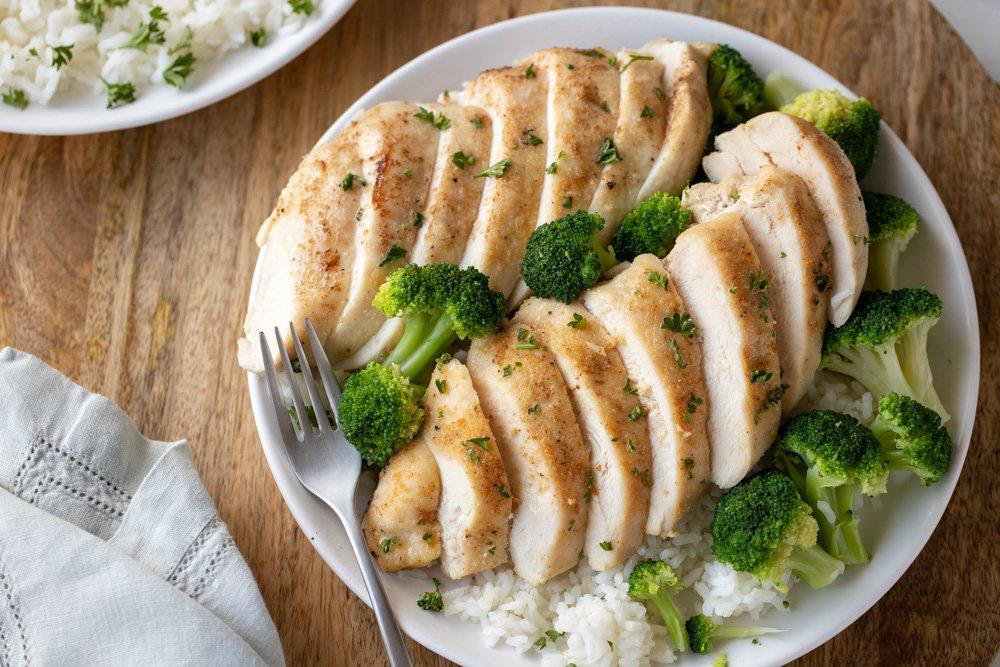 Overhead view of white plate with rice, broccoli and sliced toaster oven chicken breast. Fork and gray napkin on left.