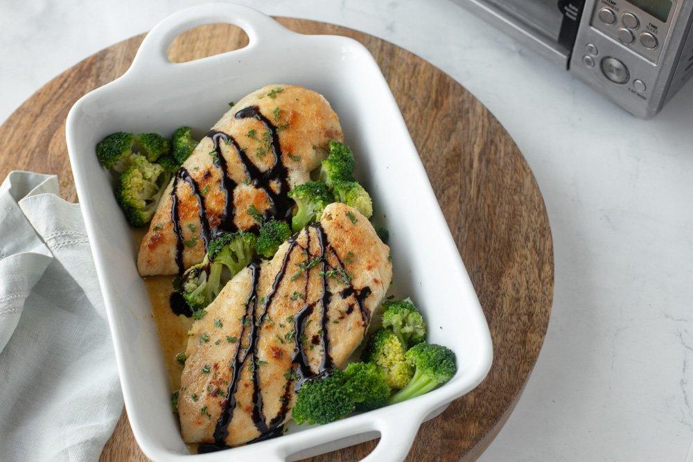 Overhead view of baked chicken breast topped with balsamic glaze and broccoli in white baking dish. Round wood board underneath. Gray napkin on left and toaster oven in upper right.
