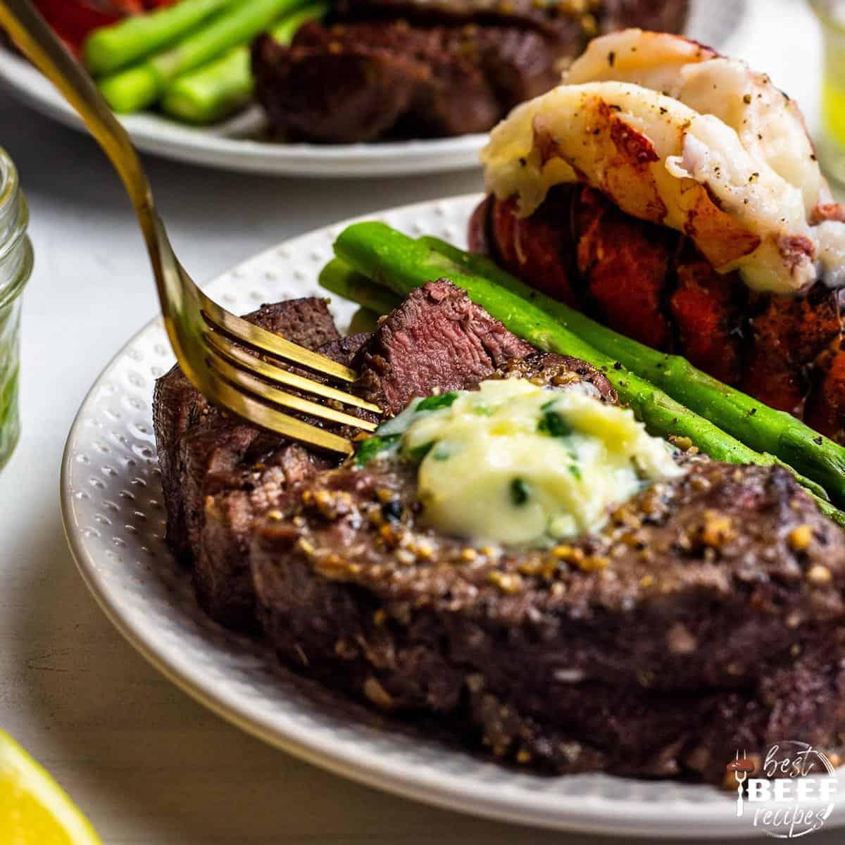 Sticking a fork into an air fried steak on a plate with a butterflied lobster tail and asparagus spears
