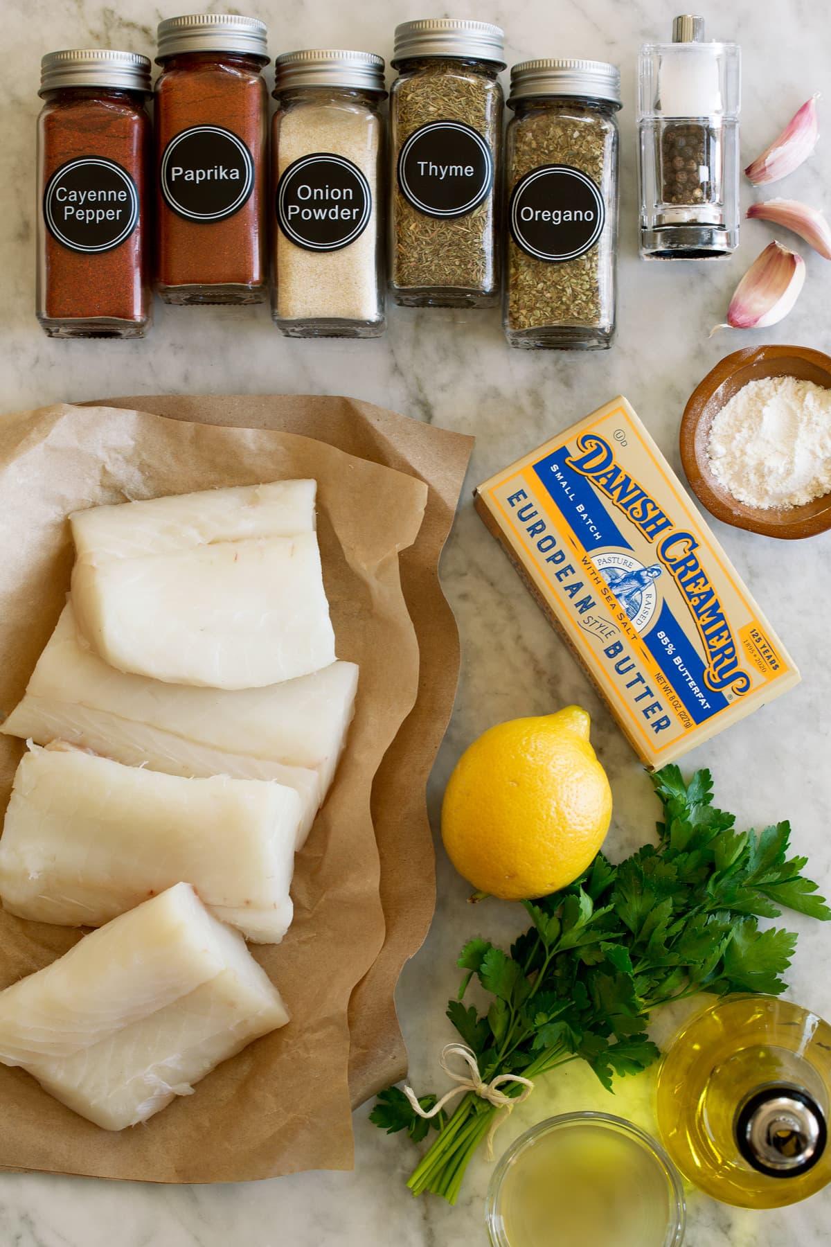 Photo of ingredients that are used to make this lemon butter cod recipe. Includes fresh cod fillets, oregano, thyme, onion powder, paprika, cayenne pepper, salt and pepper, flour, garlic, butter, lemon, parsley, olive oil, and chicken broth.