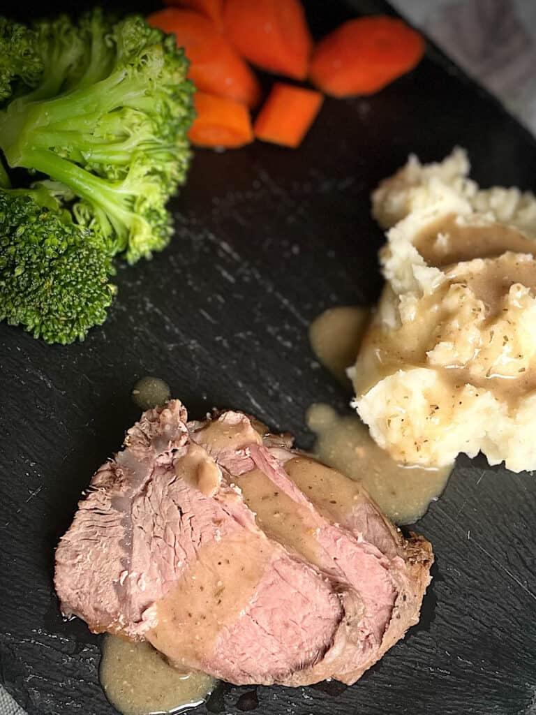 Three slices of cross rib roast sit on a black slate plate beside mashed potatoes, carrots, and broccoli. Gravy is smothering the roast and potatoes.