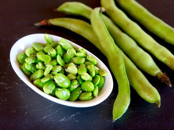 A white dish with fresh green fava beans next to 5 fava bean pods on a black background.