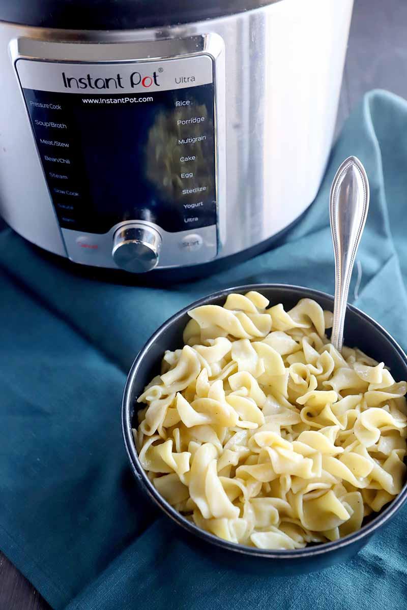 Vertical image of a bowl of pasta with a metal spoon inserted into it on top of a blue towel next to a kitchen appliance.