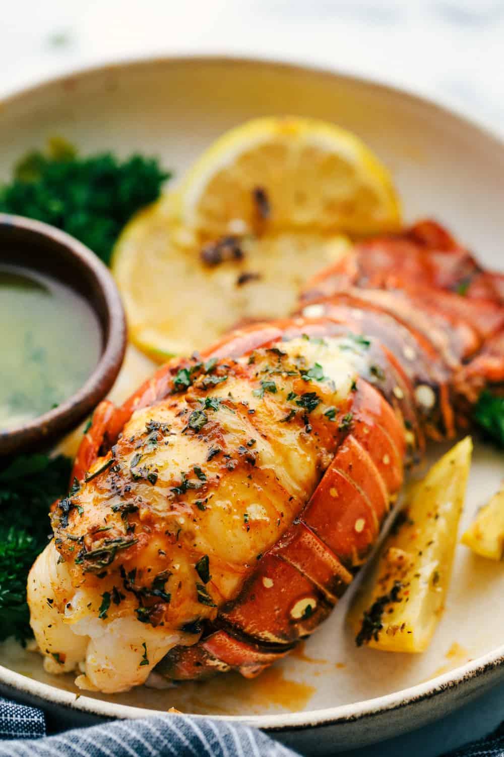 A lobster tail butterflied and served on a plate with a buttery garlic herb sauce with lemon slices.