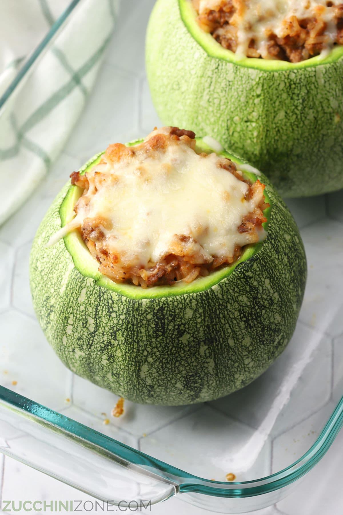 Two stuffed round zucchini with melted cheese on top in a casserole dish.