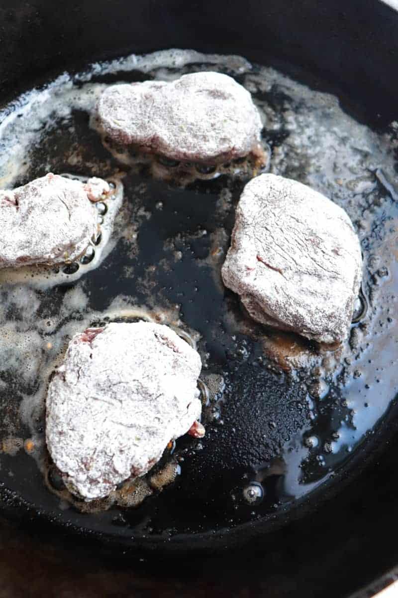 venison medallions coated in flour frying in a cast iron skillet