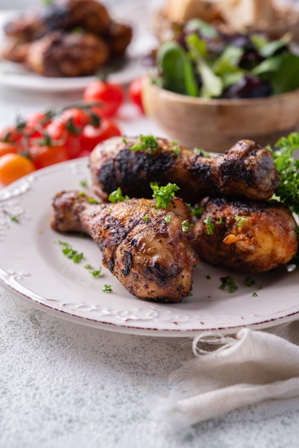 Three grilled chicken legs on a white plate, garnished with parsley. In the back is a bowl of salad, fresh cherry tomatoes, and another plate of grilled chicken legs.