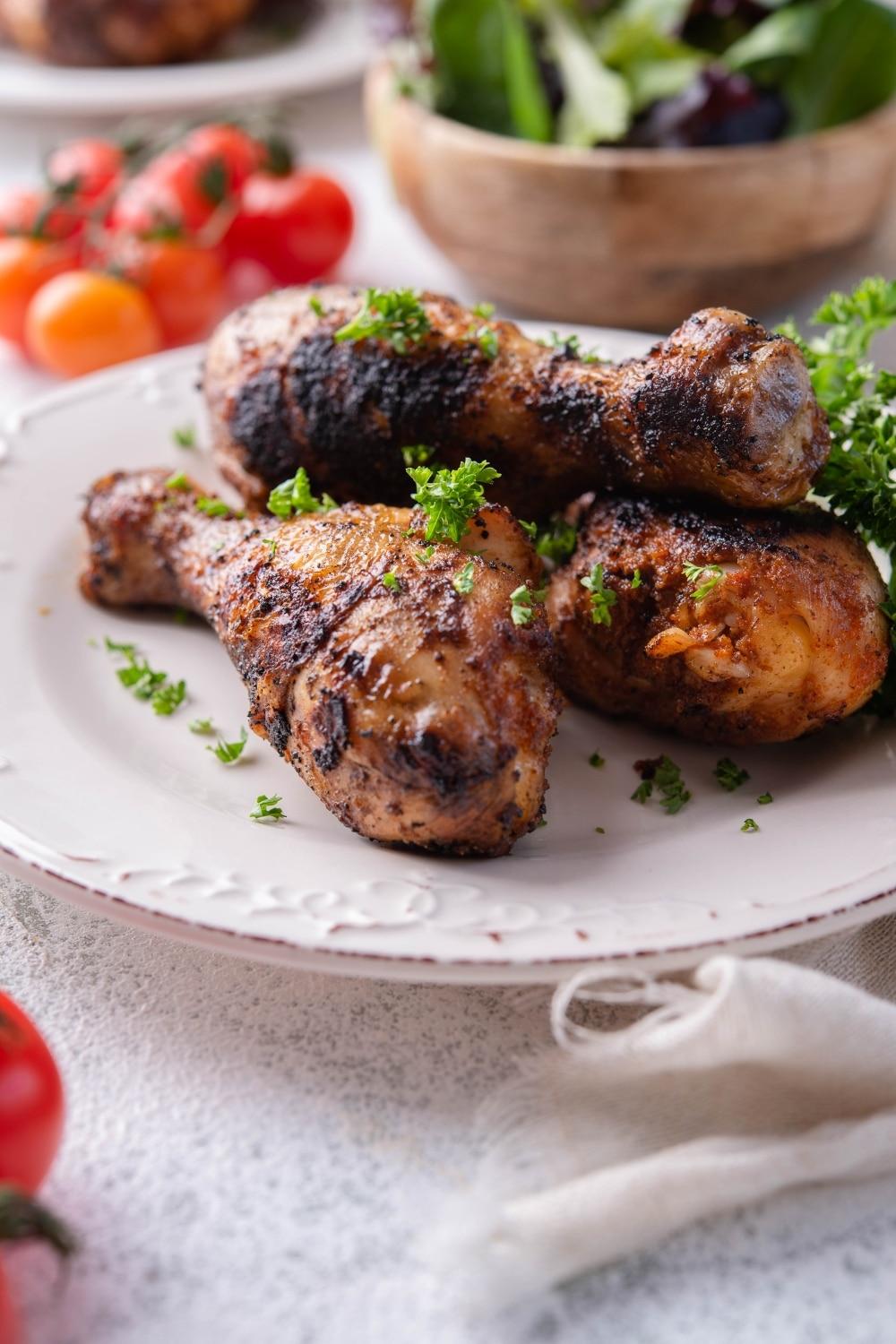 Grilled chicken legs garnished with parsley on a white plate, in front of a bowl of salad and fresh cherry tomatoes.