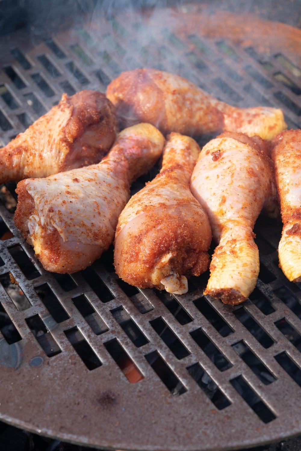 Six raw chicken drumsticks cooking on a charcoal grill.