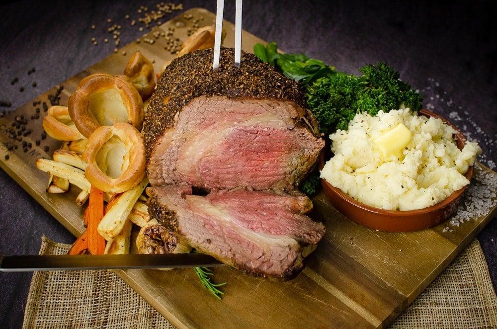 Cutting our Rolled rib of beef boneless roast joint into slices with a silver meat fork and silver knife on a wooden chopping board served with flawless Yorkshire puddings, roasted parsnips, carrots, garlic bulbs,sprigs of rosemary, broccoli and creamy mash in a brown bowl