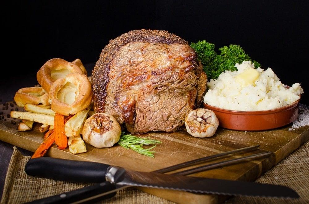 Rolled rib of beef boneless roast joint served on a wooden chopping board with flawless Yorkshire puddings, roasted parsnips, carrots, garlic bulbs,sprigs of rosemary, broccoli and creamy mash in a brown bowl