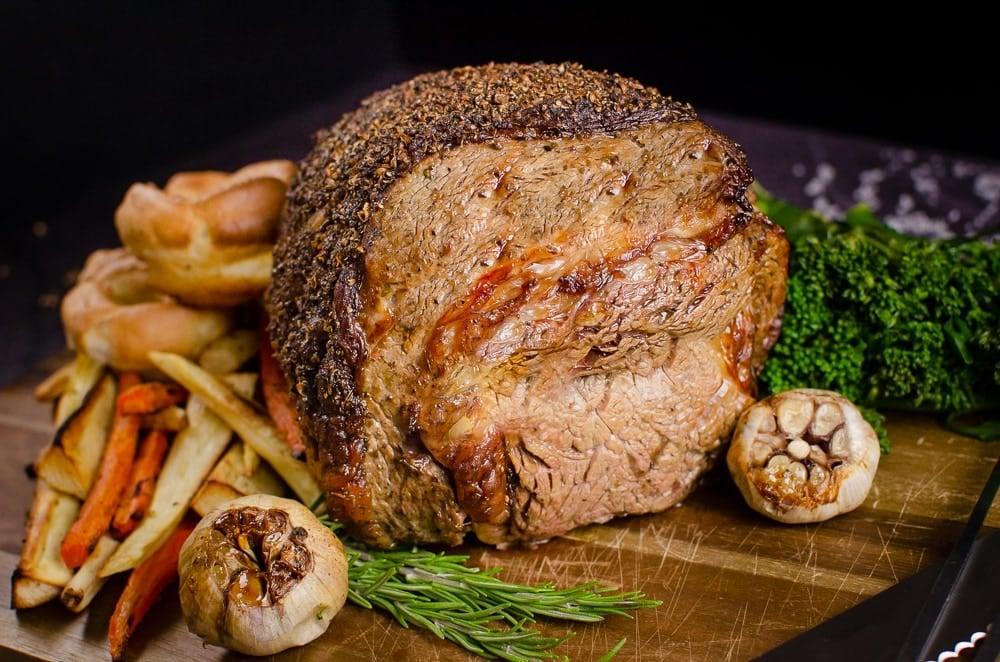 Rolled rib of beef boneless roast joint served on a wooden chopping board with flawless Yorkshire puddings, roasted parsnips, carrots, garlic bulbs,sprigs of rosemary and broccoli