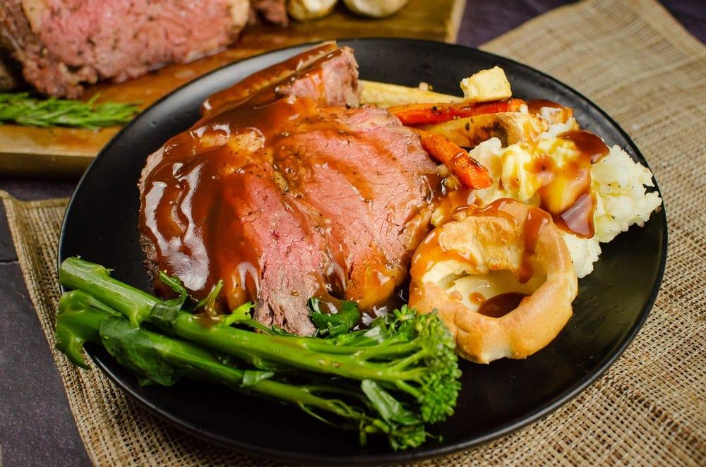 Slices of Rolled rib of beef boneless roast joint on a wooden chopping board served with flawless Yorkshire puddings, roasted parsnips, carrots, garlic bulbs,sprigs of rosemary, broccoli and creamy mash in a brown bowl