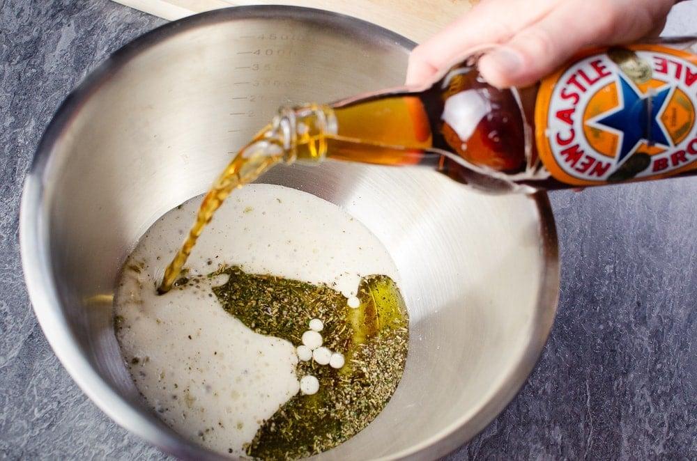 Newcastle Brown Ale being poured on top of the olive oil and mixed herbs in a silver bowl to make our beef marinade