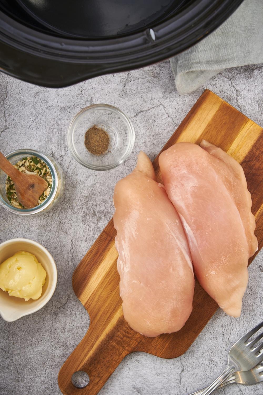 An assortment of ingredients including two raw chicken breasts on a wooden cutting board and small bowls of salt and pepper, a seasoning blend, and butter.