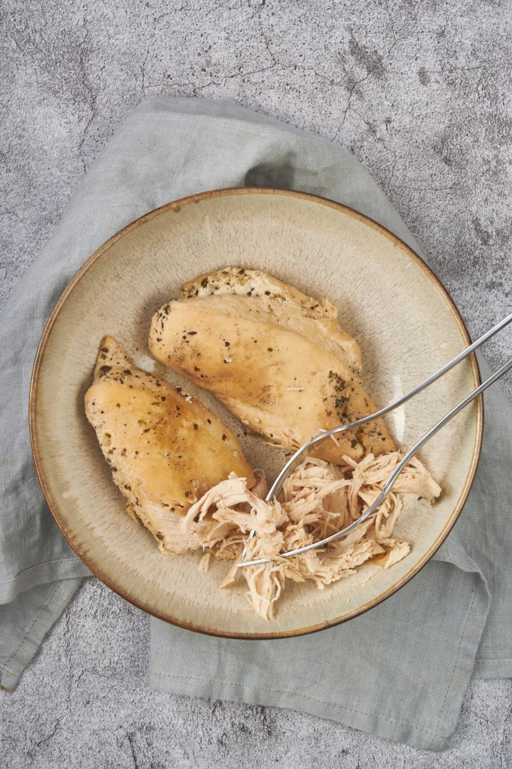 Two seasoned chicken breasts in a bowl with one chicken breast partly shredded using two forks.