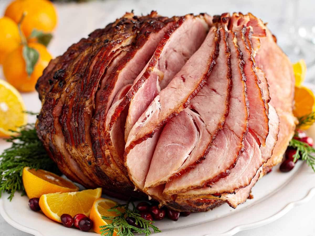 A spiral-sliced glazed ham on a white plate garnished with orange slices, cranberries, and sprigs of evergreen.