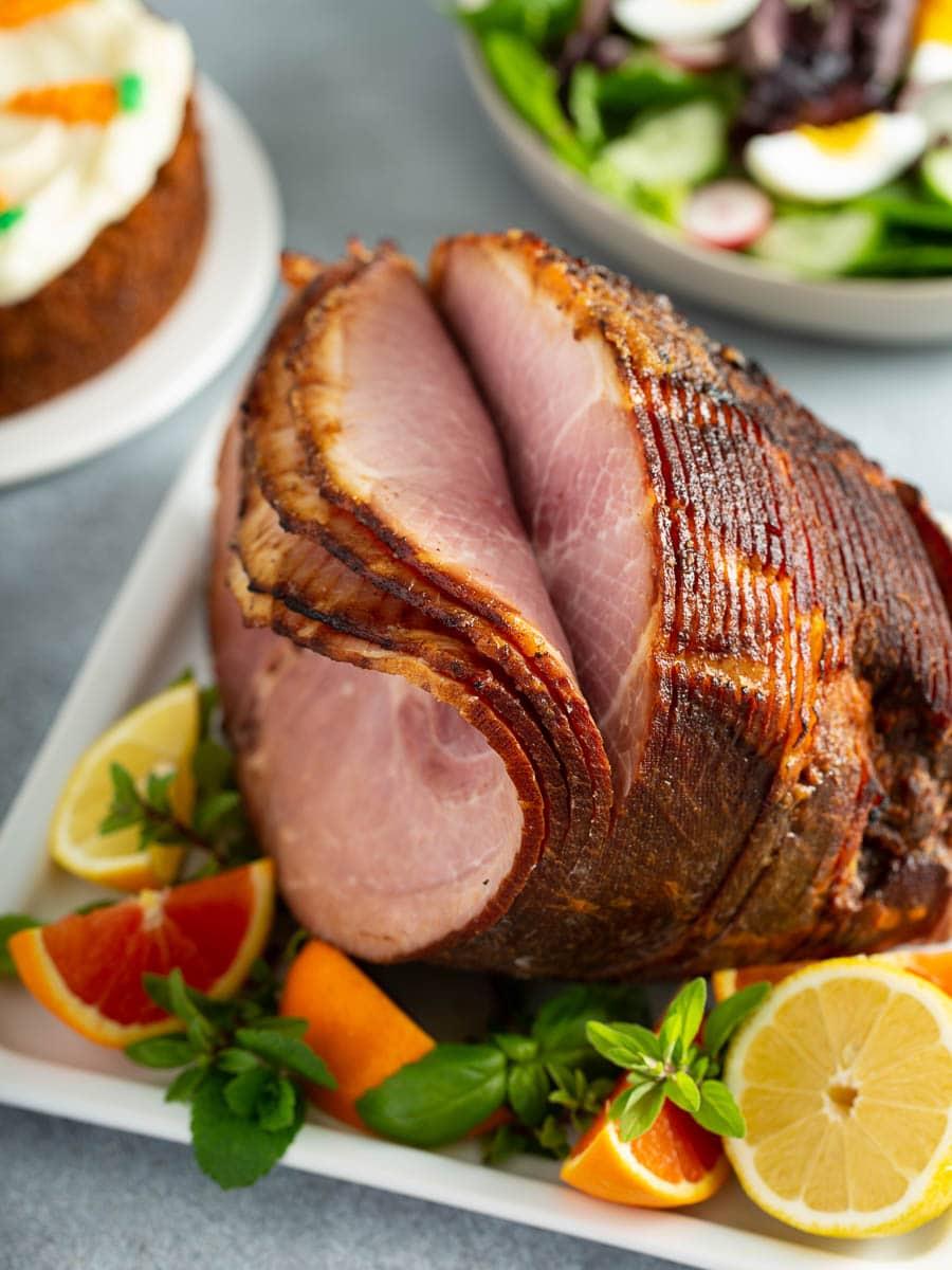 A sliced ham on a white plate, garnished with lemon, lime, and orange slices, with a salad and cake in the background.