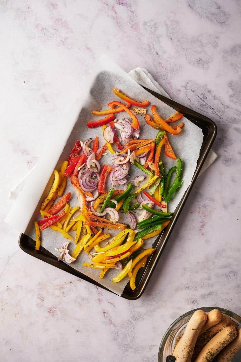 Sliced bell peppers and onions seasoned with herbs on a parchment paper lined baking tray.