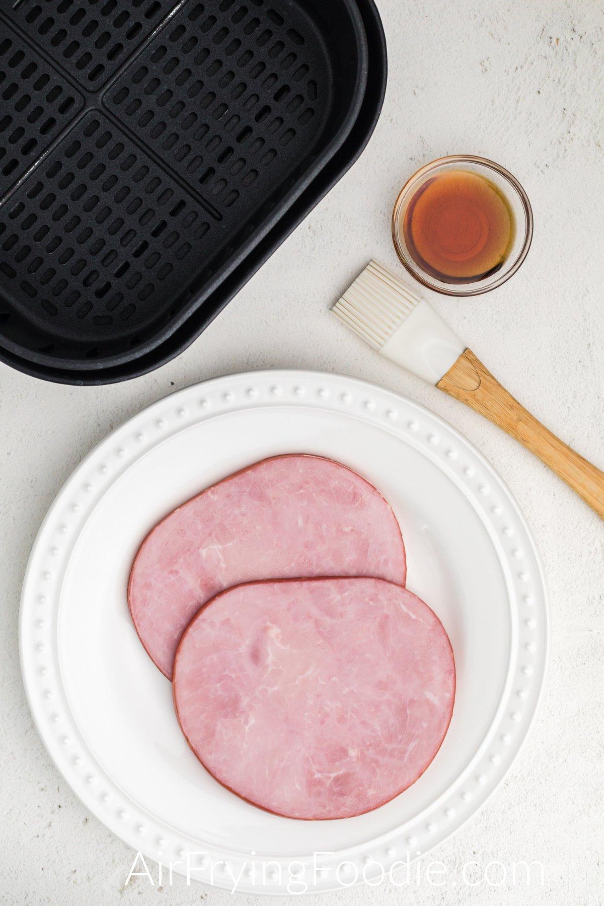 Ham steaks and maple syrup with a silicone basting brush on a white table.