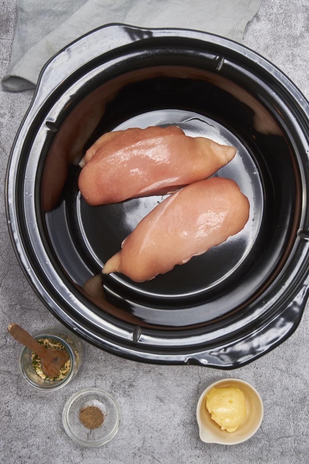 Two raw chicken breasts in the bottom of a black crock pot next to a small bowl of butter, a small bowl of salt and pepper, and a small bowl of seasonings.