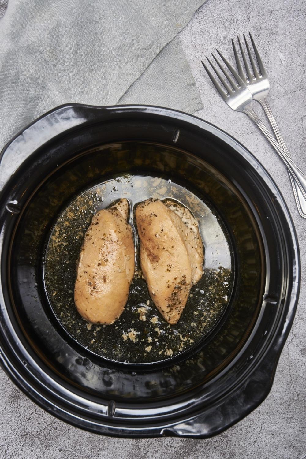 Two seasoned and cooked chicken breasts in the bottom of a black crockpot.