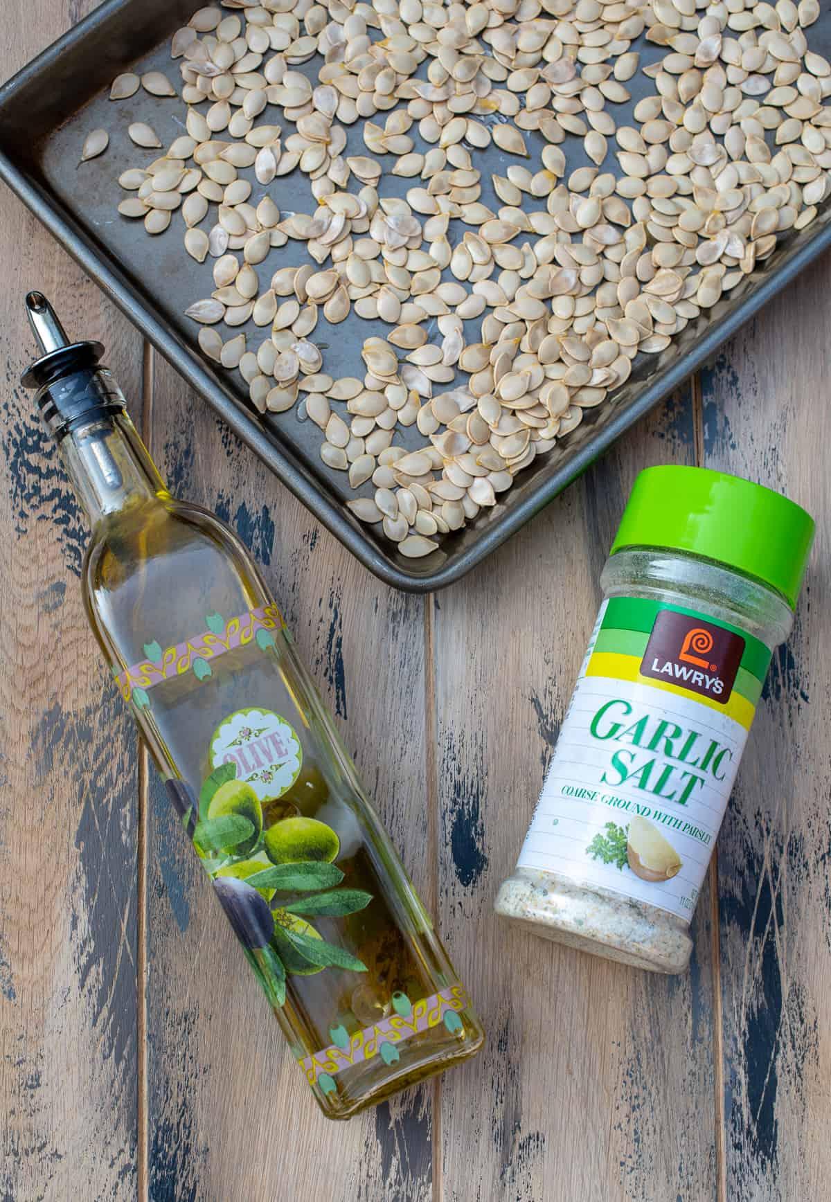 A top down shot of acorn squash seeds on a baking sheet, a glass bottle of olive oil, and a container of garlic salt on a brown board.