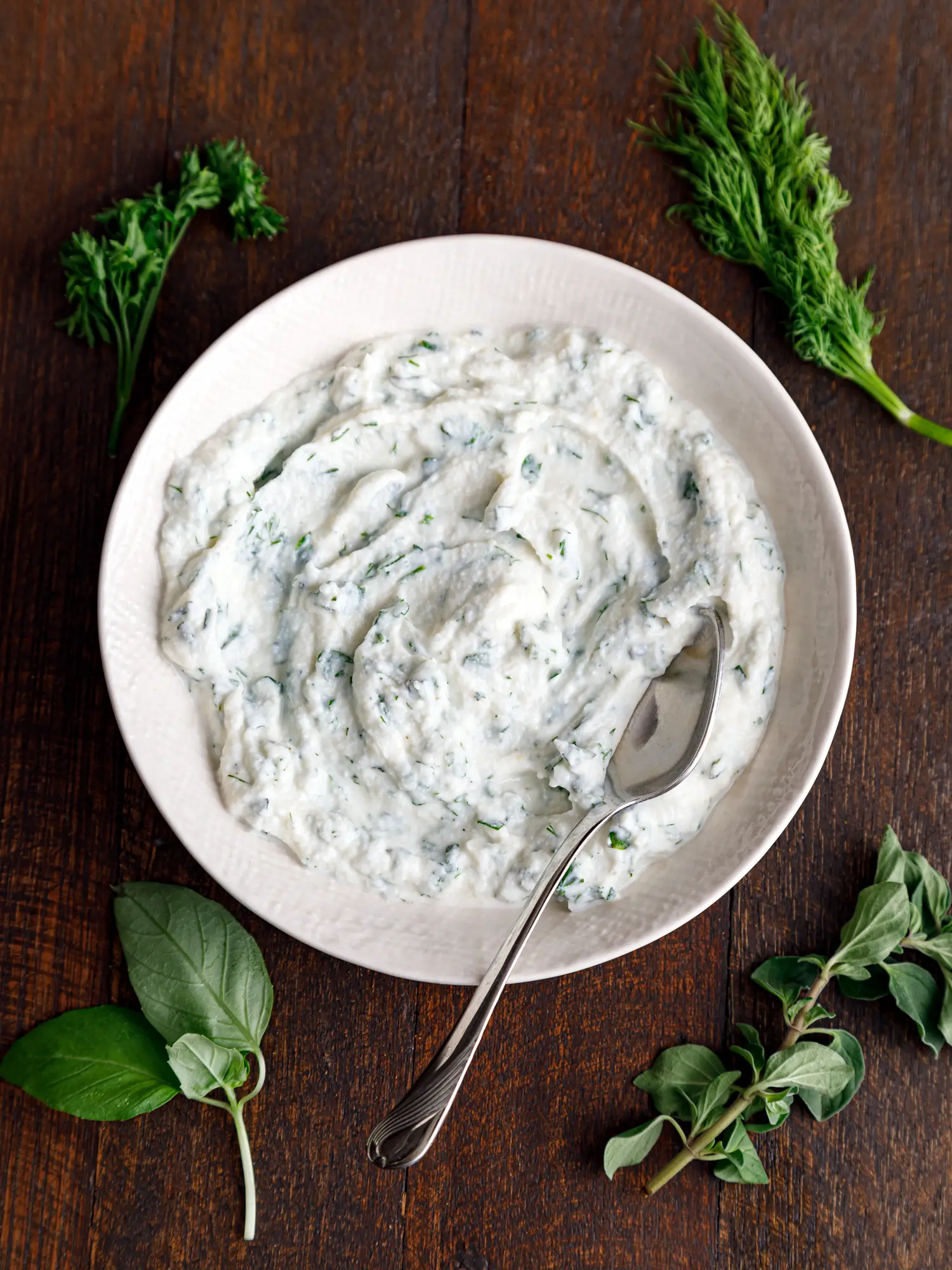 A white bowl of herbed ricotta cheese on a table surrounded by fresh herbs like basil, dill, and parsley