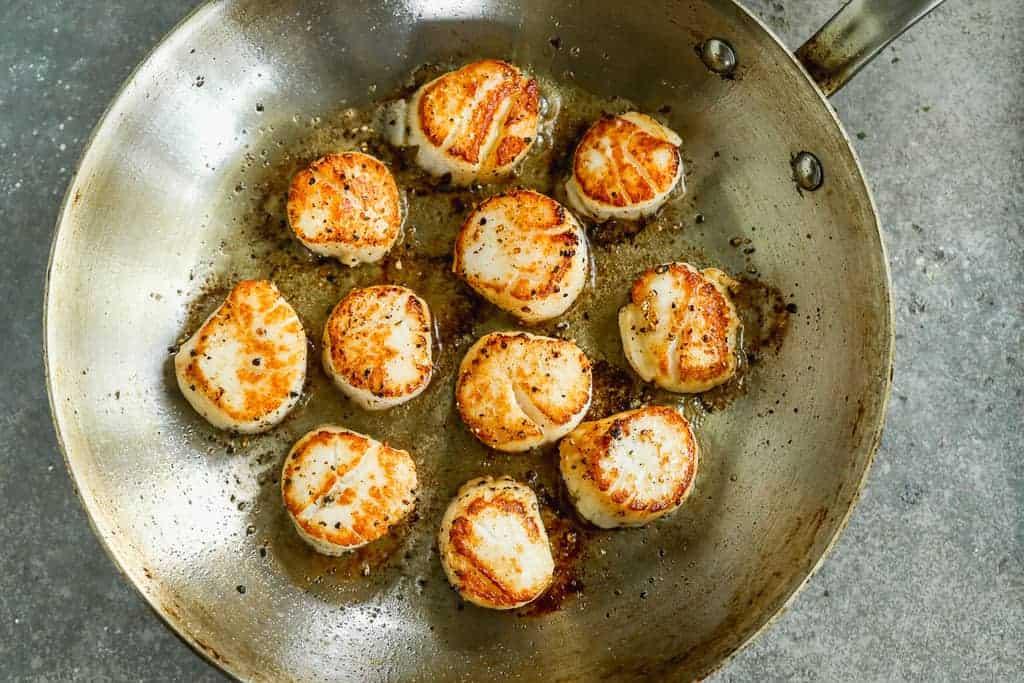 Scallops searing in a pan, flipped once to show golden crust.