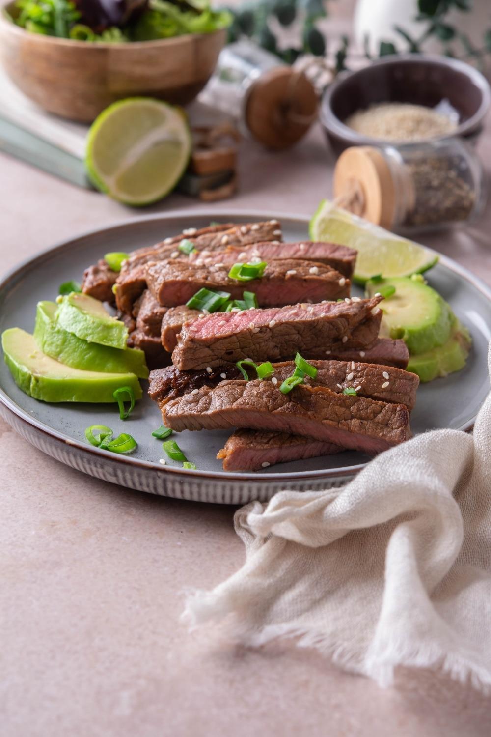 Sliced sirloin tip steak on a plate served with avocado slices and garnished with lime wedges and chopped green onion.