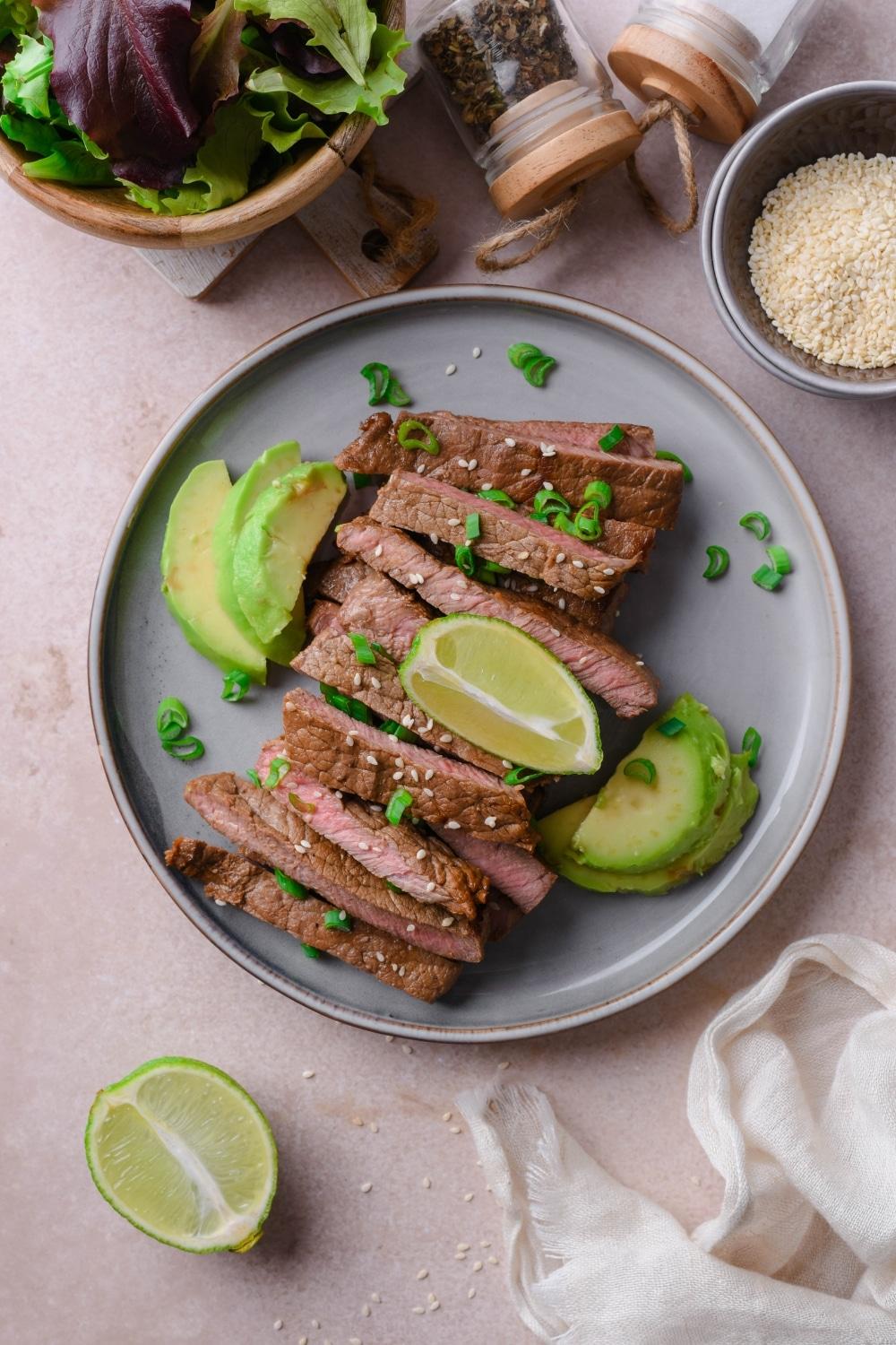 Sliced medium sirloin tip steak on a plate served with avocado slices and garnished with lime wedges and chopped green onion.