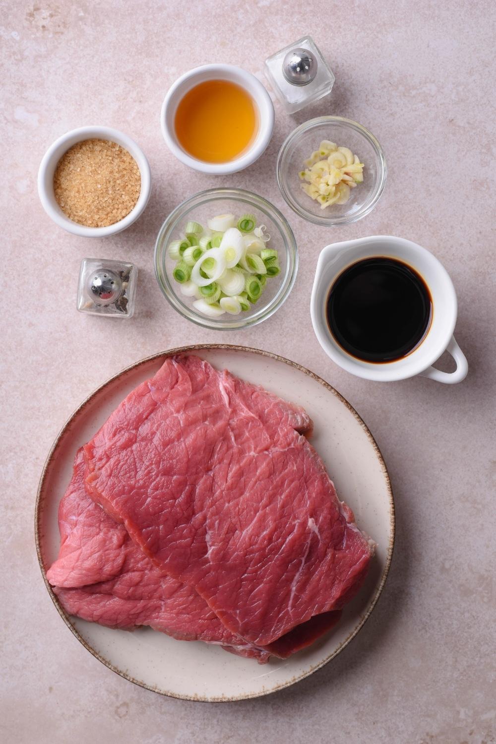 Raw sirloin tip steak on a plate, with small bowls of soy sauce, rice vinegar, brown sugar, sliced garlic cloves, sliced green onion, and salt and pepper shakers.