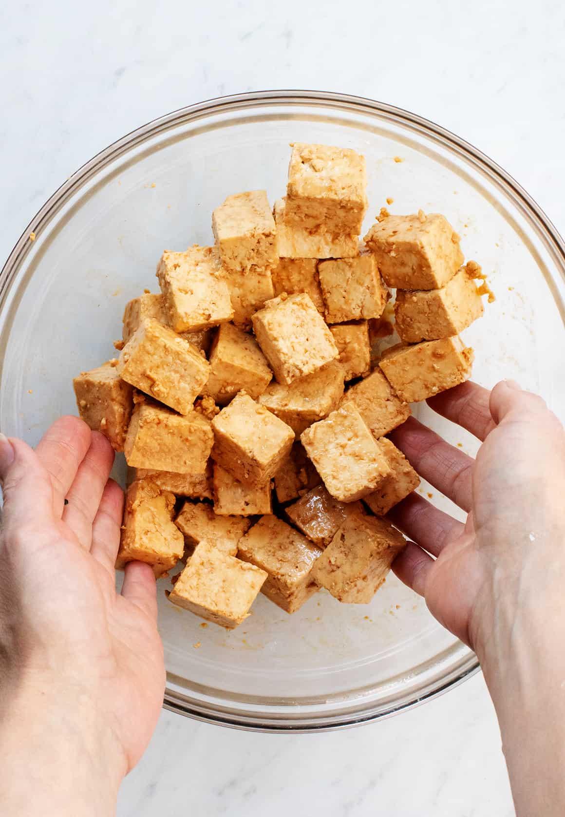 Hands tossing cubed tofu with seasonings in glass bowl